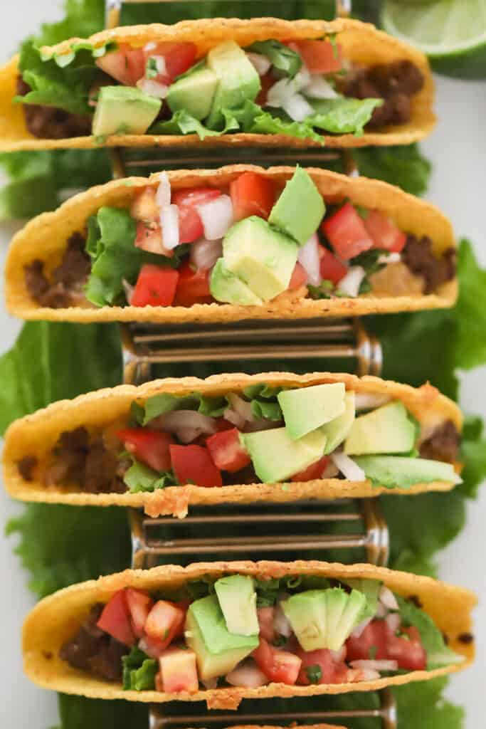 Ground Beef Tacos resting in a taco holder, topped with tomatoes, lettuce and avocados. taco recipe ground beef.  how to make tacos, crunchy tacos, how to make ground beef for tacos, taco crispy