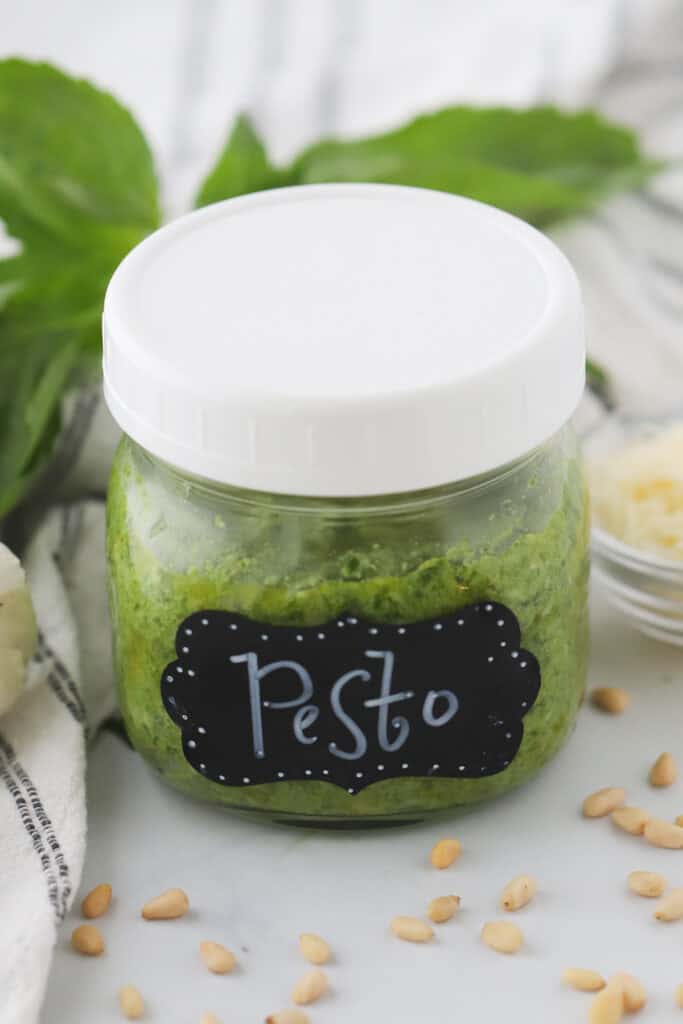 Homemade Pesto in a small jar on a table surrounded by pine nuts and basil. This recipe shows you how to make fresh pesto at home.
