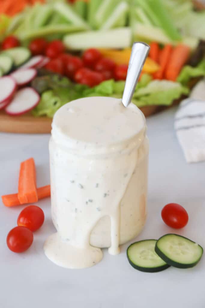 Homemade Ranch Dressing overflowing out of a glass jar, with a serving platter full of vegetables in the background. Ranch salad dressing recipe.