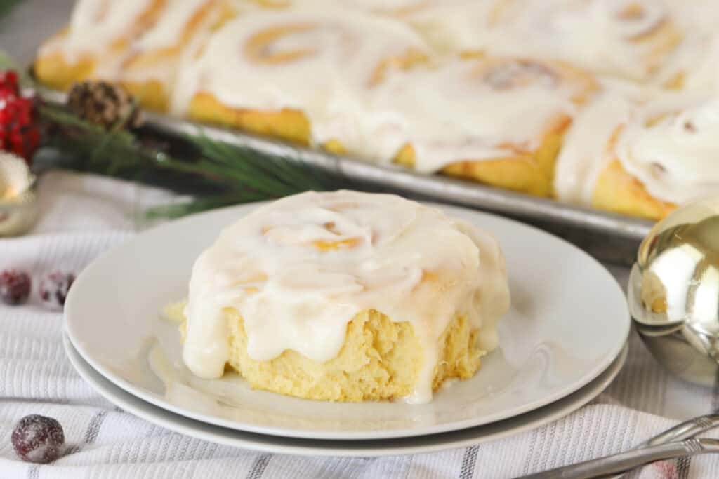 A small white plate with a Lemon Sweet Roll covered in frosting and a full pan of sweet rolls in the background. lemon butter icing, cindy cinnamon rolls. 