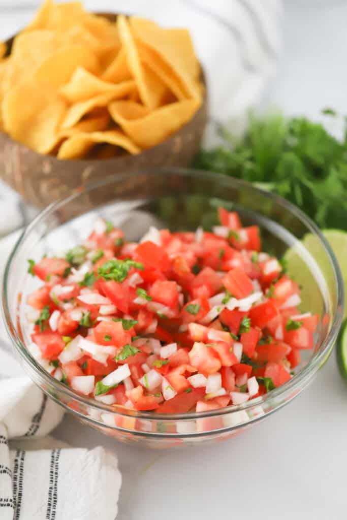 A glass serving bowl full of fresh Pico de Gallo, with chips, fresh limes and cilantro in the background.