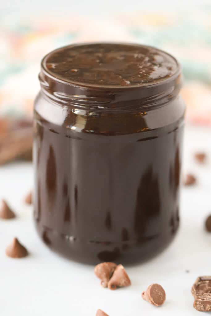 A glass jar full of chocolate sauce recipe on a table surrounded by mini chocolate chips, chocolate syrup for milk.