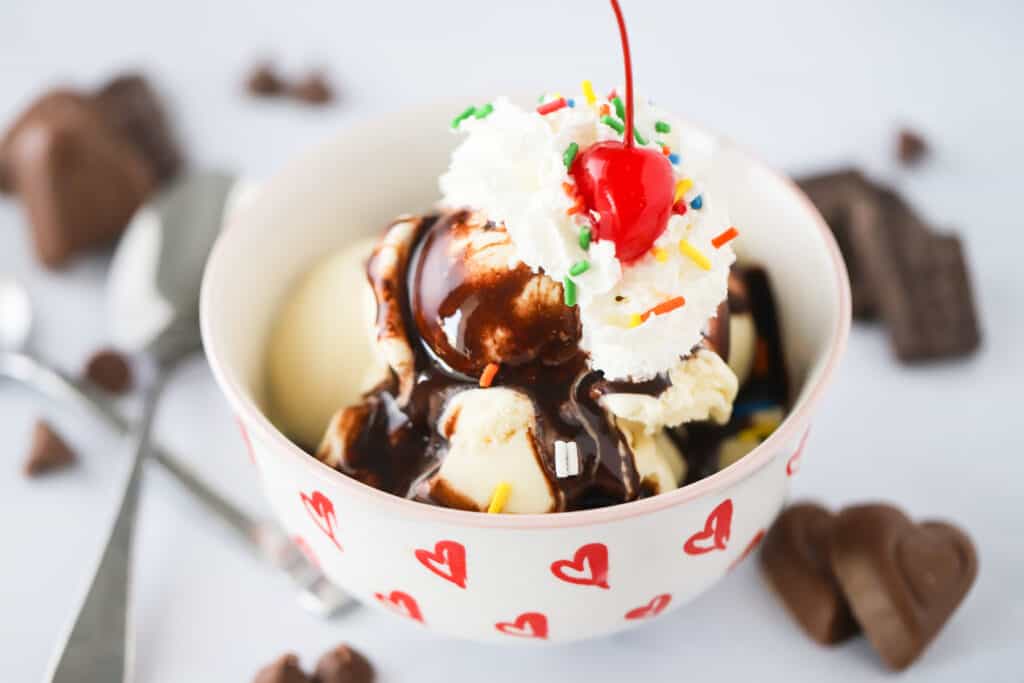A white serving bowl full of ice cream drizzled with Homemade Chocolate Syrup and topped with whipped cream and a cherry.