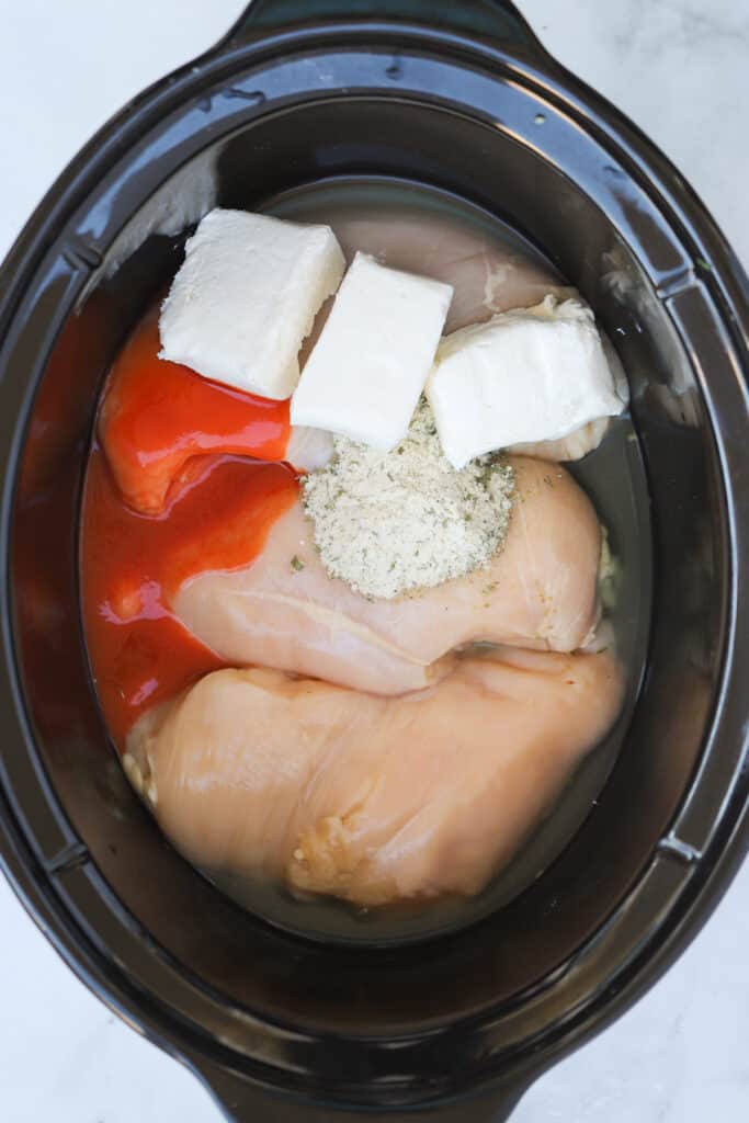 Raw chicken, cream cheese, hot sauce, broth and seasoning inside of a slow cooker. ingredients for buffalo chicken crockpot recipe.