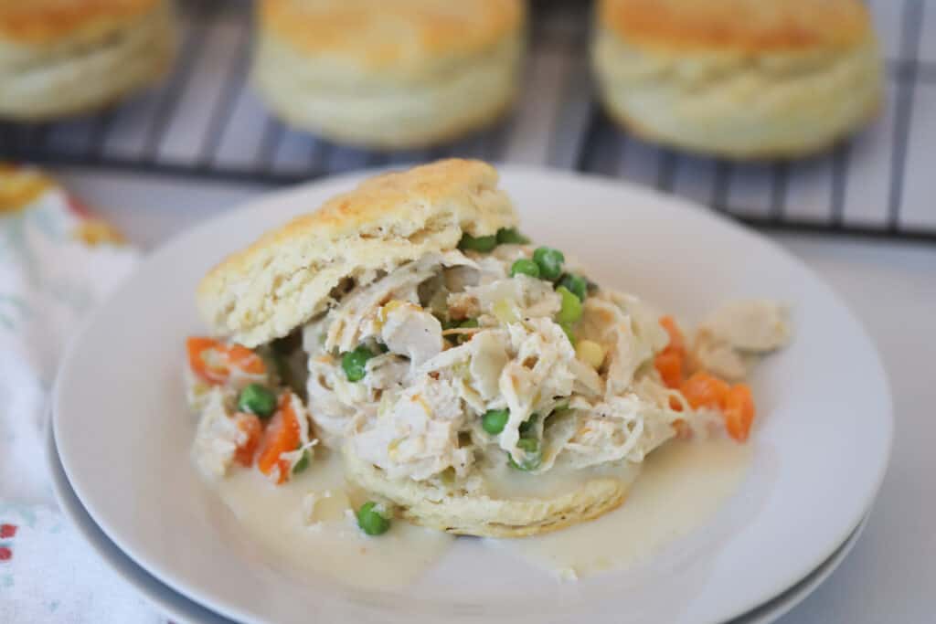 A white serving plate with chicken pot pie in crock pot recipe served over a sliced biscuit. chicken pot pie in slow cooker. slow cooker chicken pot pie with biscuits.