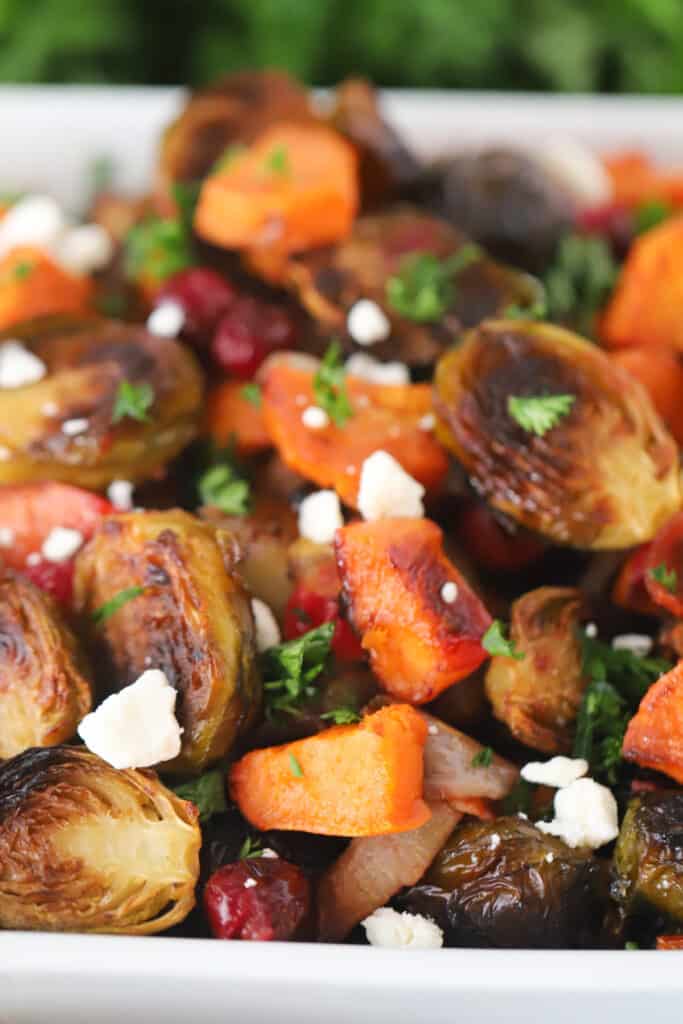 A close up of oven-roasted brussels sprouts and sweet potatoes in a baking dish, topped with feta cheese crumbles and fresh parsley.