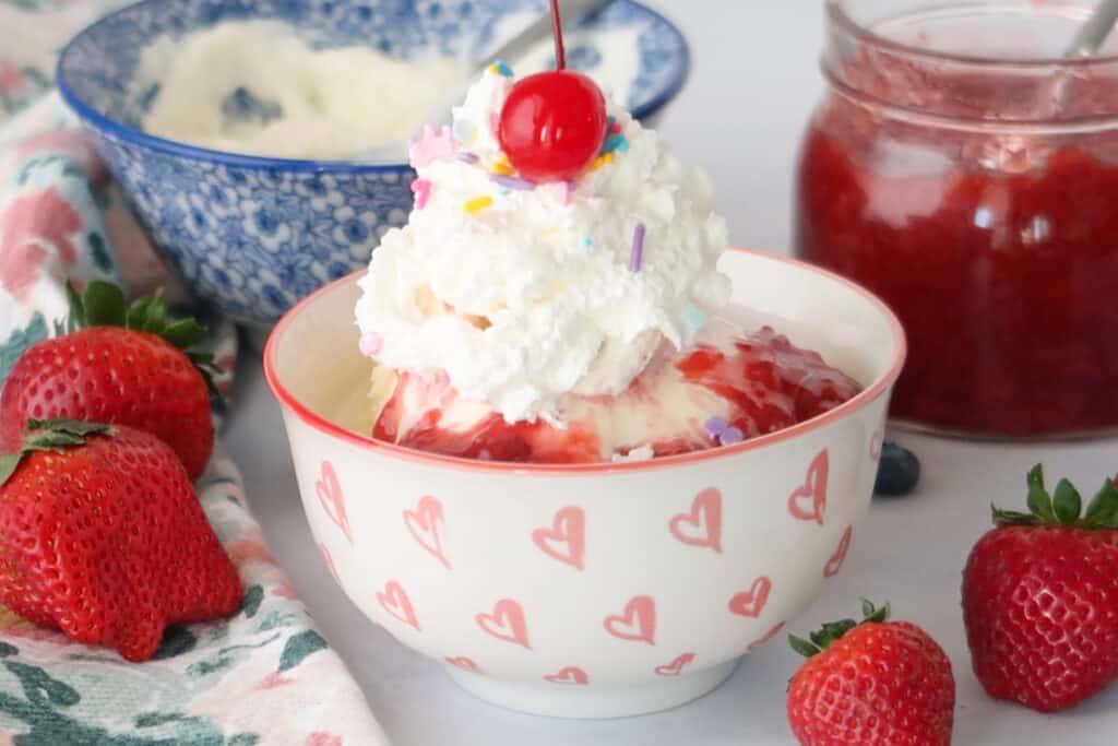 A serving bowl full of ice cream, strawberry sauce and whipped cream.