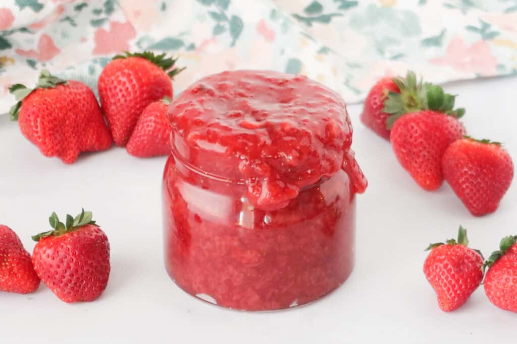 A glass jar overflowing with strawberry compote on a table surrounded by fresh strawberries, strawberry sauce recipes.