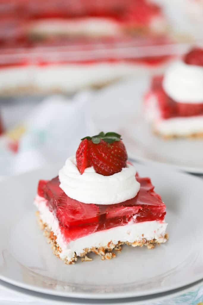 A white serving place with a slice of jello pretzel salad topped with whipped cream and a fresh strawberry, strawberry jello salad.
