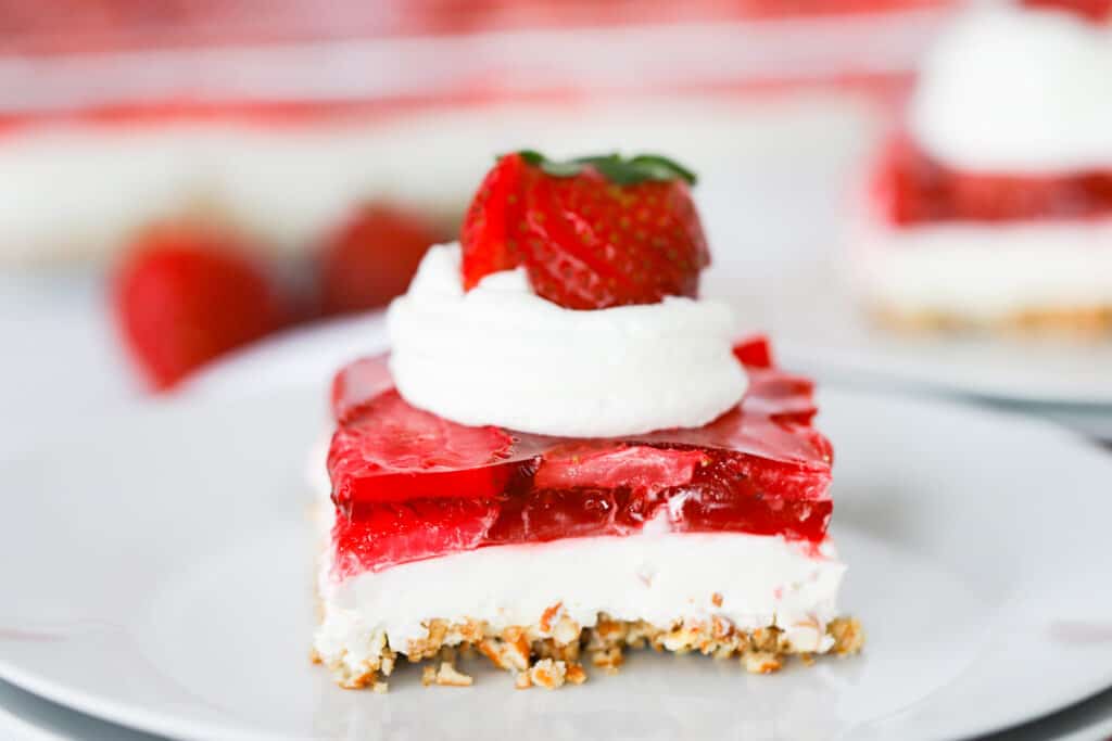 A white serving plate with a square of pretzel salad recipe strawberry topped with whipped cream and fresh berries, a strawberry salad dessert recipe. 