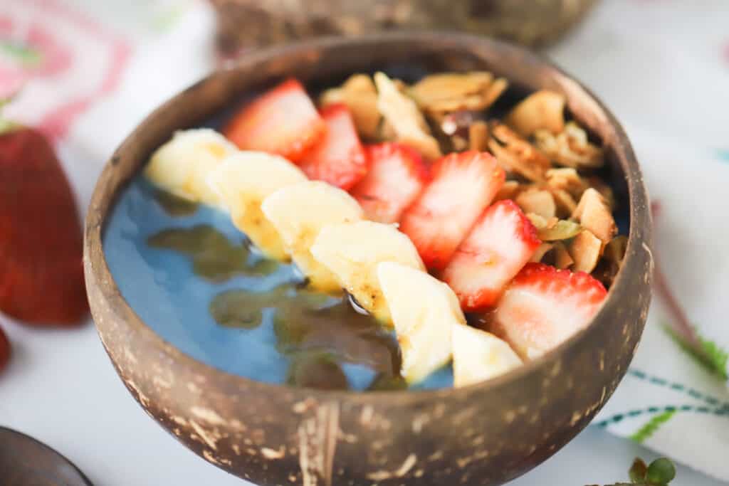 Blue spirulina smoothie bowl topped with honey, bananas, strawberries and granola on a table.