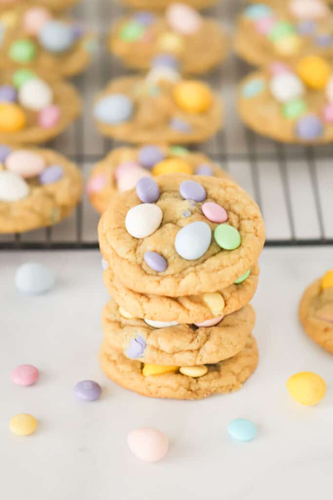 A stack of Cadbury Mini Egg Cookies on a table with a cooling rack full of more cookies in the background. cute Cadbury cookies recipe.