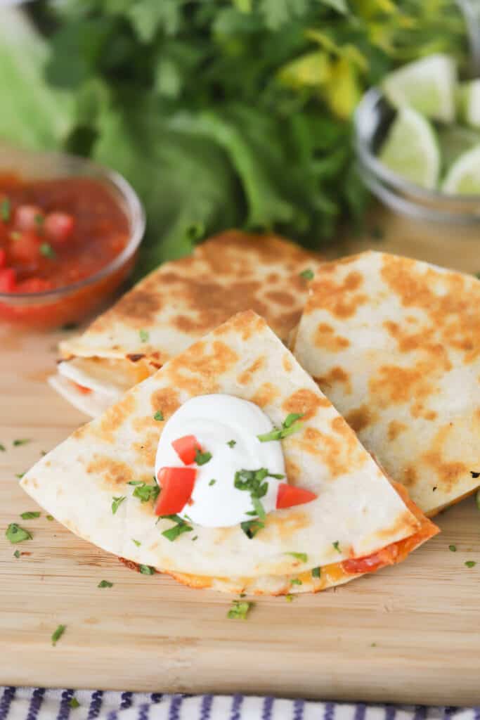 A tabletop with crispy Chicken Quesadillas cut into quarters and topped with sour cream, tomatoes and fresh cilantro. Simple chicken quesadilla recipe.