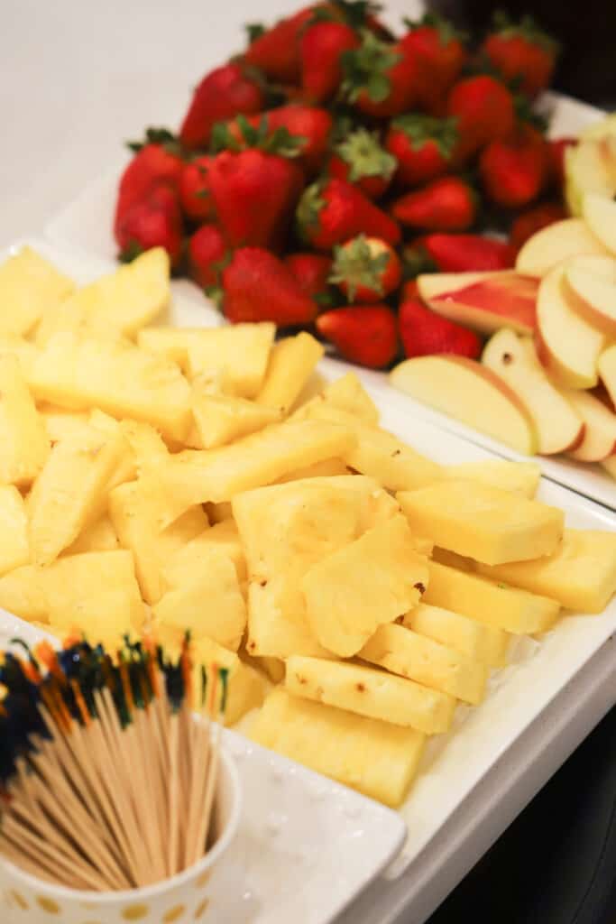 Dipping ideas on a table including fresh pineapple wedges, whole strawberries, apples slices and a cup of toothpicks.