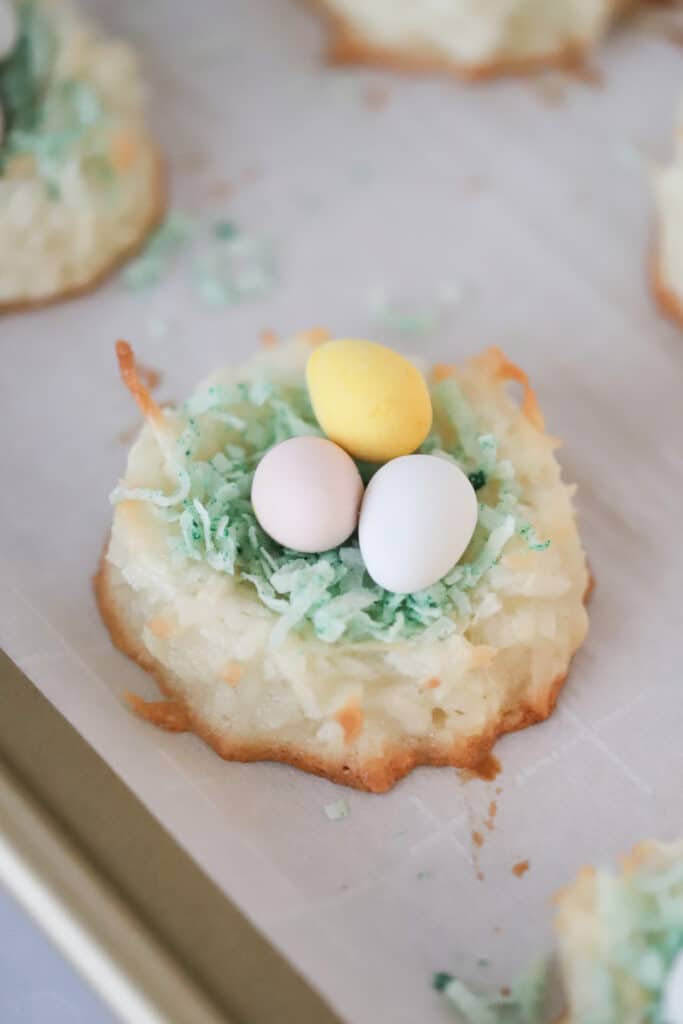 Coconut Macaroon Easter Nests topped with green colored shredded coconut and candy eggs on a baking sheet.