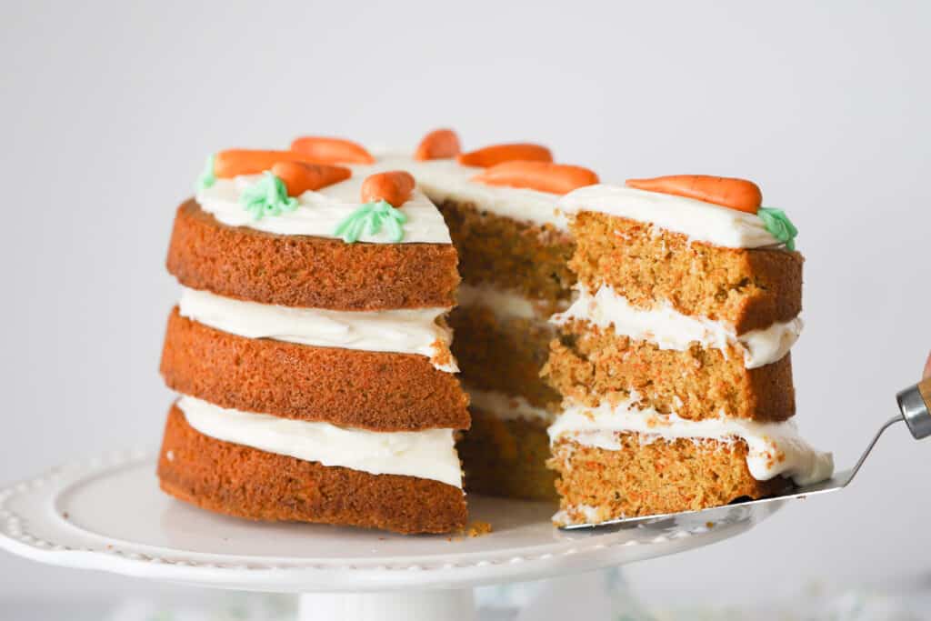 A three-tiered Carrot Cake with cream cheese frosting on top of the cake and in between the layers.  A slice is being removed using a cake server.
