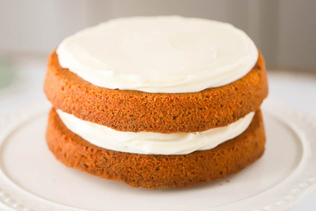 A side view of layers of Carrot Cake on a white plate with cream cheese frosting in between and on top.