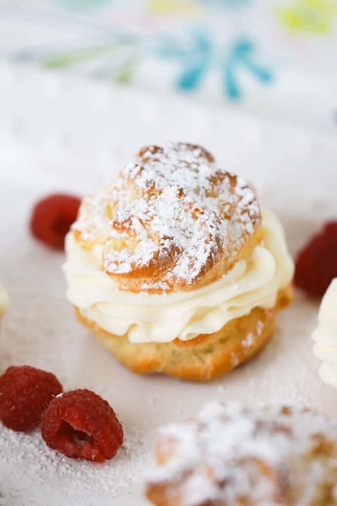 A cream puff recipe filled with custard and topped with powdered sugar on a table next to fresh raspberries. 