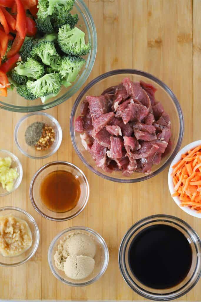 Raw ingredients in small glass bowls and ramekins full of raw steak, cut vegetables and ingredients to make stir fry sauce.