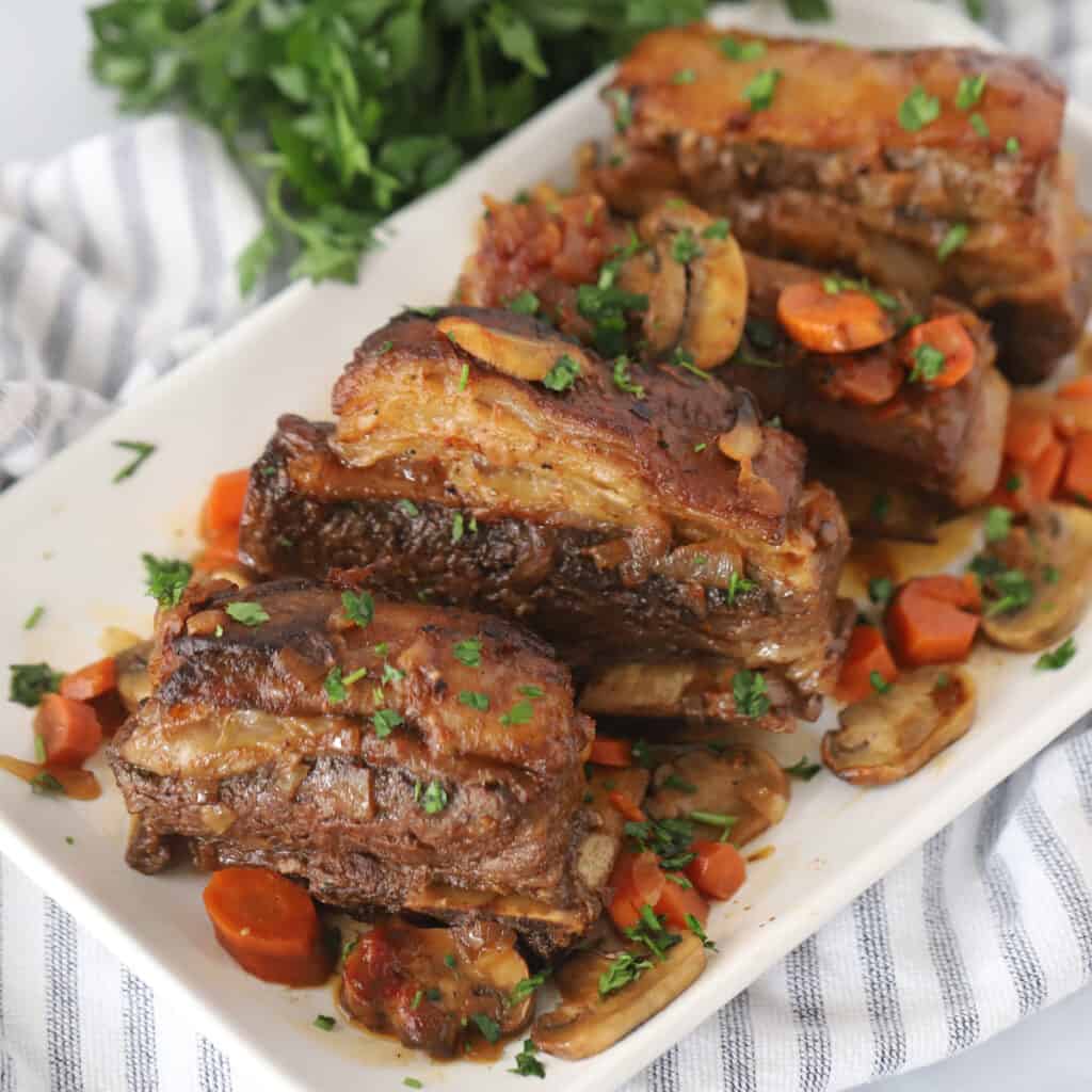 Beef short ribs on a large serving platter with carrots, mushrooms, and fresh parsley. Valentine’s day dinner at home. valentines dinner recipes