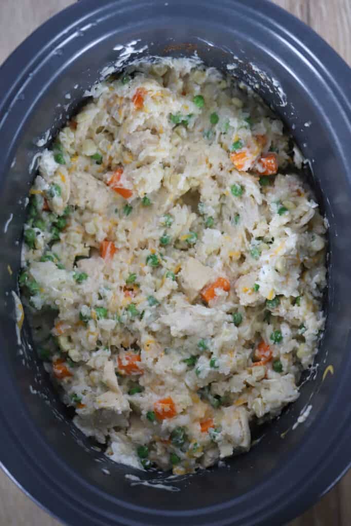 A slow cooker filled with cooked rice, chicken, peas, carrots and cheese.