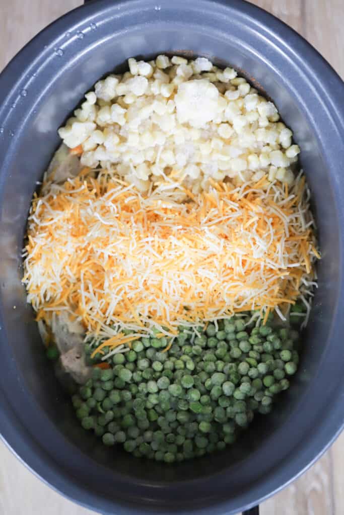 A crockpot from above showing peas, shredded cheese and corn added over the top of Cheesy Chicken and Rice.
