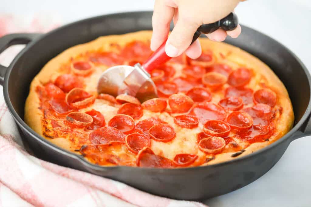 A pepperoni Deep Dish Pizza baked in a cast iron skillet and being cut using a pizza cutter.