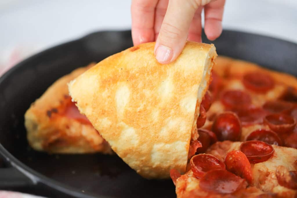 A hand grabbing a slice of pepperoni pizza from a cast iron skillet.