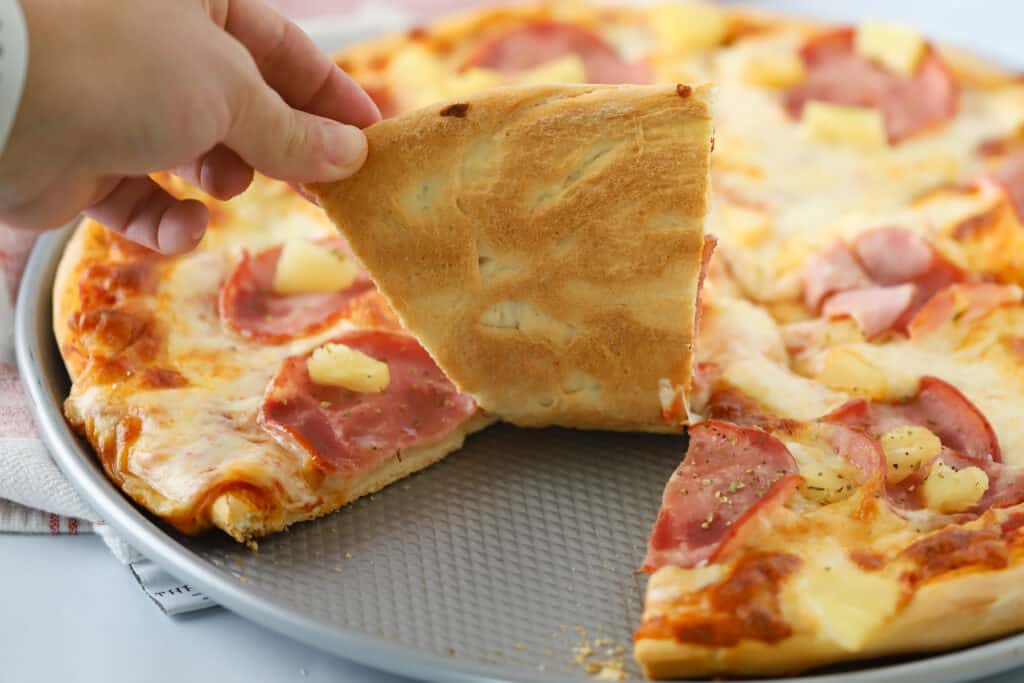 A hand grabbing a slice of Hawaii pizza from a pizza tray. recipe for Hawaiian Pizza.