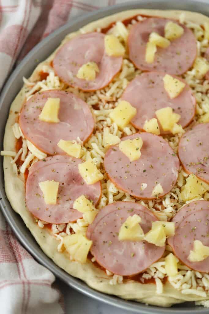 Pizza dough on a pizza pan topped with Canadian bacon, shredded cheese, fresh pineapple and seasoning.