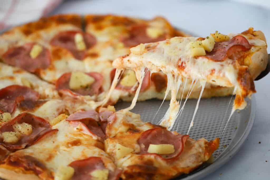 A sliced Hawaii pizza on a baking sheet with a slice being removed by a spatula. How to. make a hawaiian pizza.