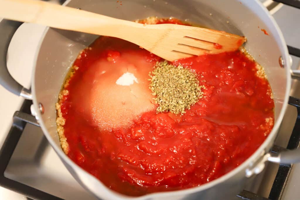 A pot with crushed tomatoes, sugar, dried herbs and a wooden spatula ready to stir.