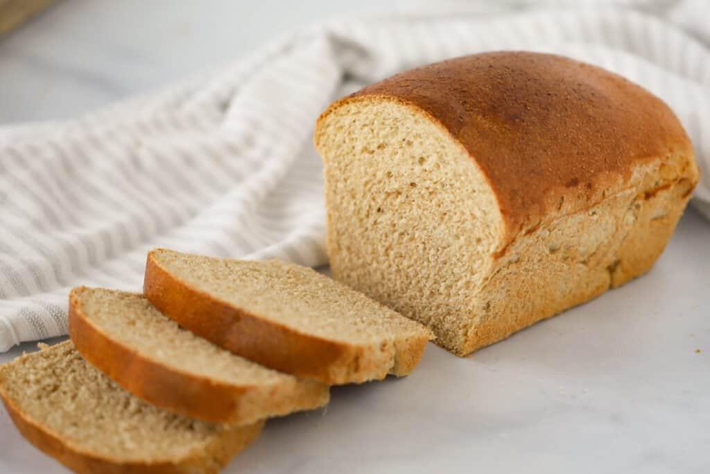 A loaf of homemade wheat bread on a table with a few slices cut from the loaf and resting flat. Soft whole wheat bread recipe.