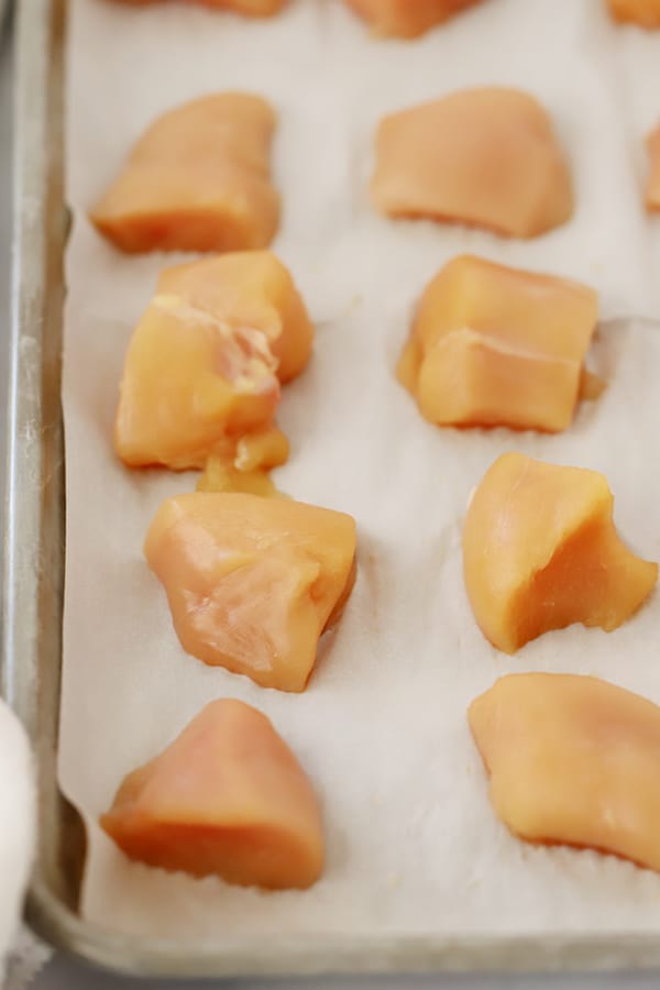 A sheet tray lined with parchment paper filled with small pieces of chicken. baked buffalo chicken breast cut into bites. Oven baked chicken bites.
