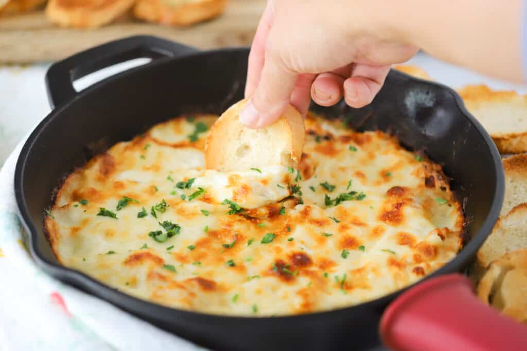 A cast iron skillet full of baked Alfredo dip, with a hand dipping a slice of bread into it.