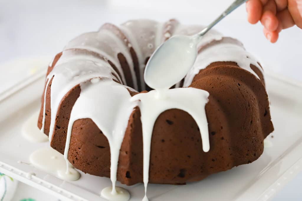 A full chocolate bundt cake on a serving plate with glaze being poured over the top using a spoon.