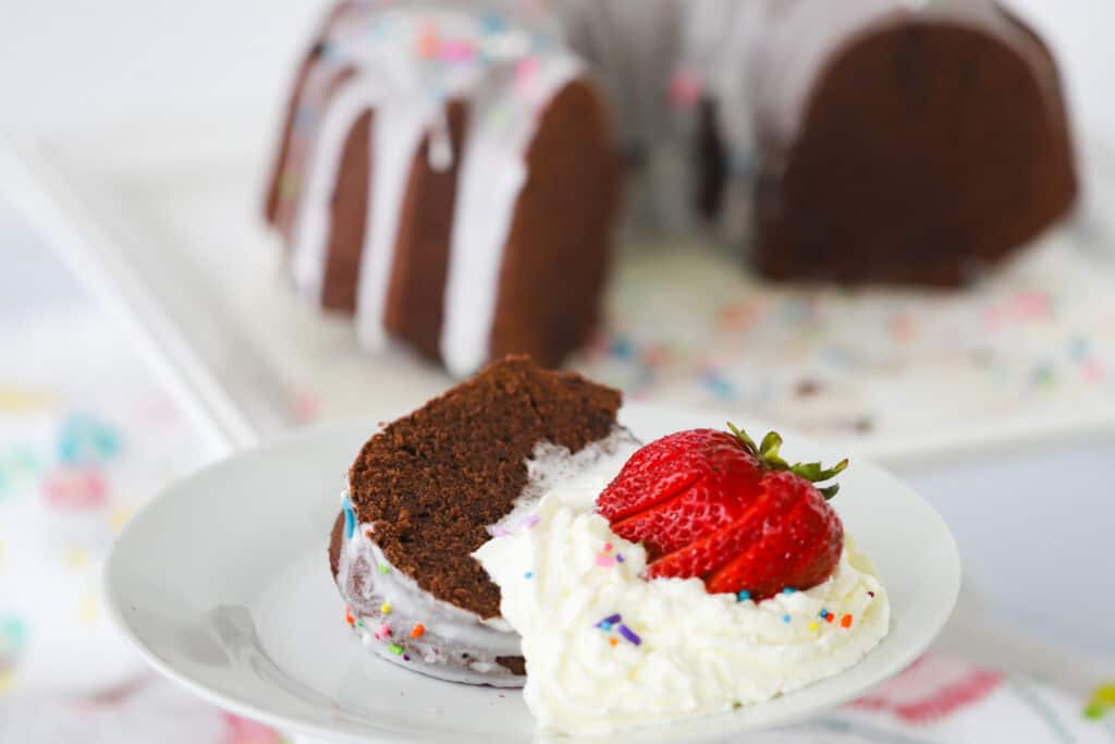A slice of chocolate pound cake on a white serving plate topped with glaze, sprinkles, whipped cream and a fresh strawberry.
