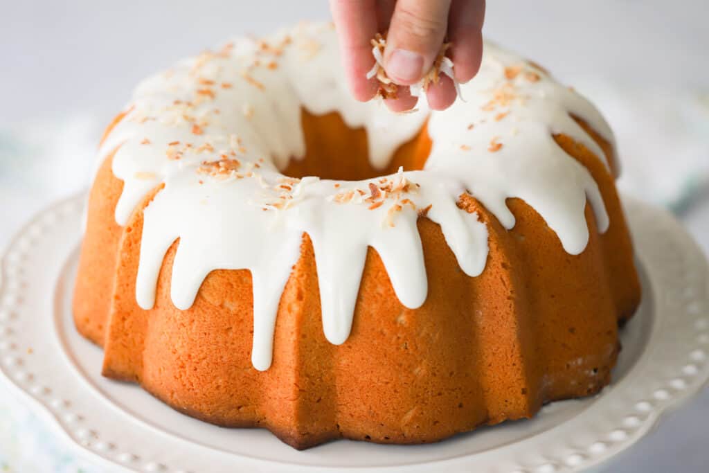 A hand sprinkling toasted coconut over a glazed bundt cake on a white serving plate. cream cheese pound cake with coconut. 