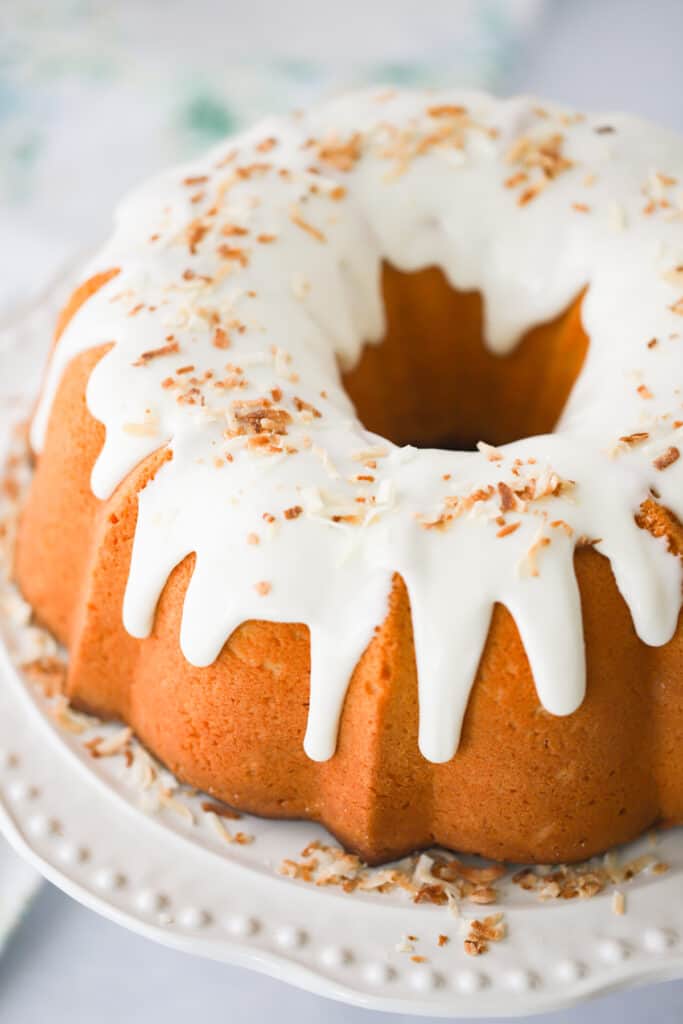 A serving plate with a large bundt cake topped with white glaze and toasted coconut.