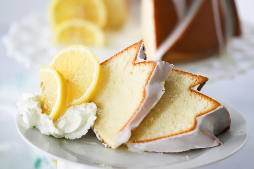 Slices of lemon pound cake on a white plate with fresh lemon and whipped cream.
