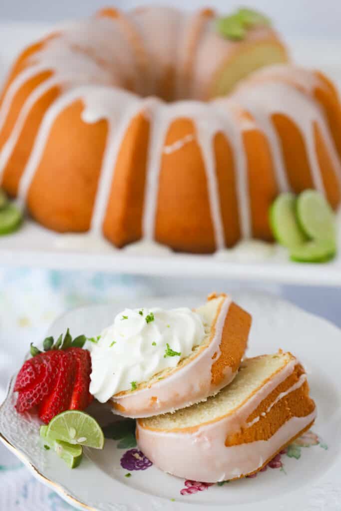 A serving plate with two slices of glazed Key Lime Pound Cake, topped with whipped cream and fresh strawberries.  A full pound cake is in the background on a cake stand. recipe for key lime pound cake. 