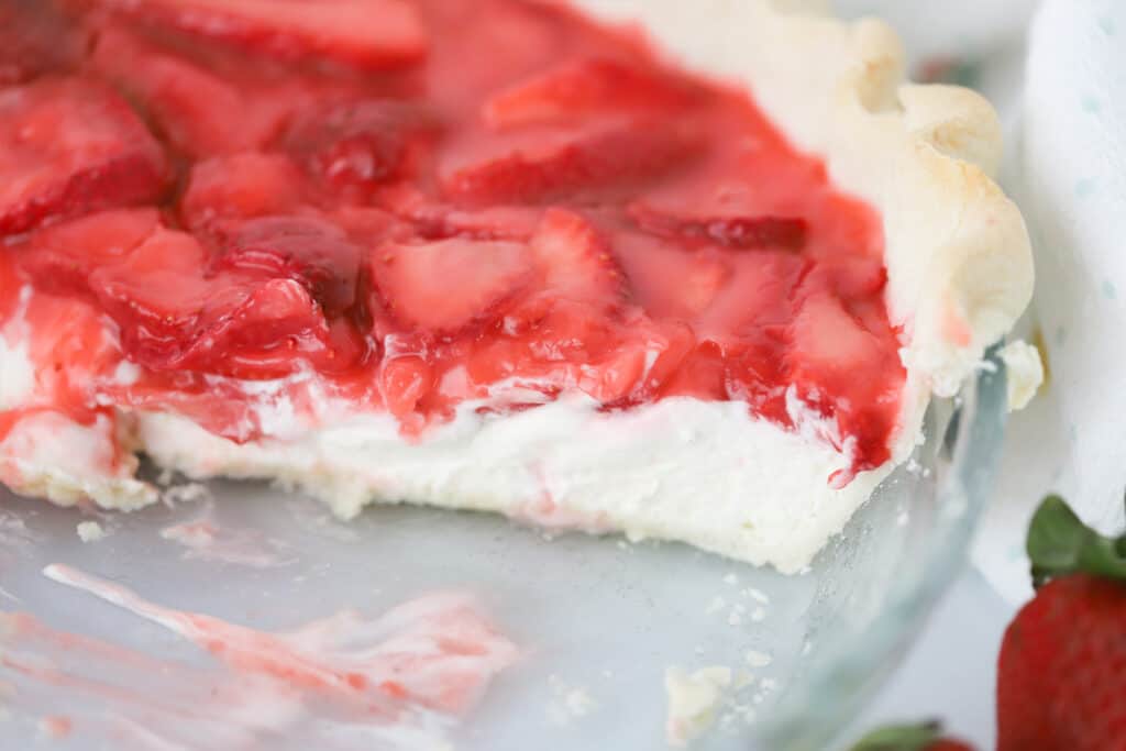A pie dish with a strawberry and cream pie that has a few slices removed.