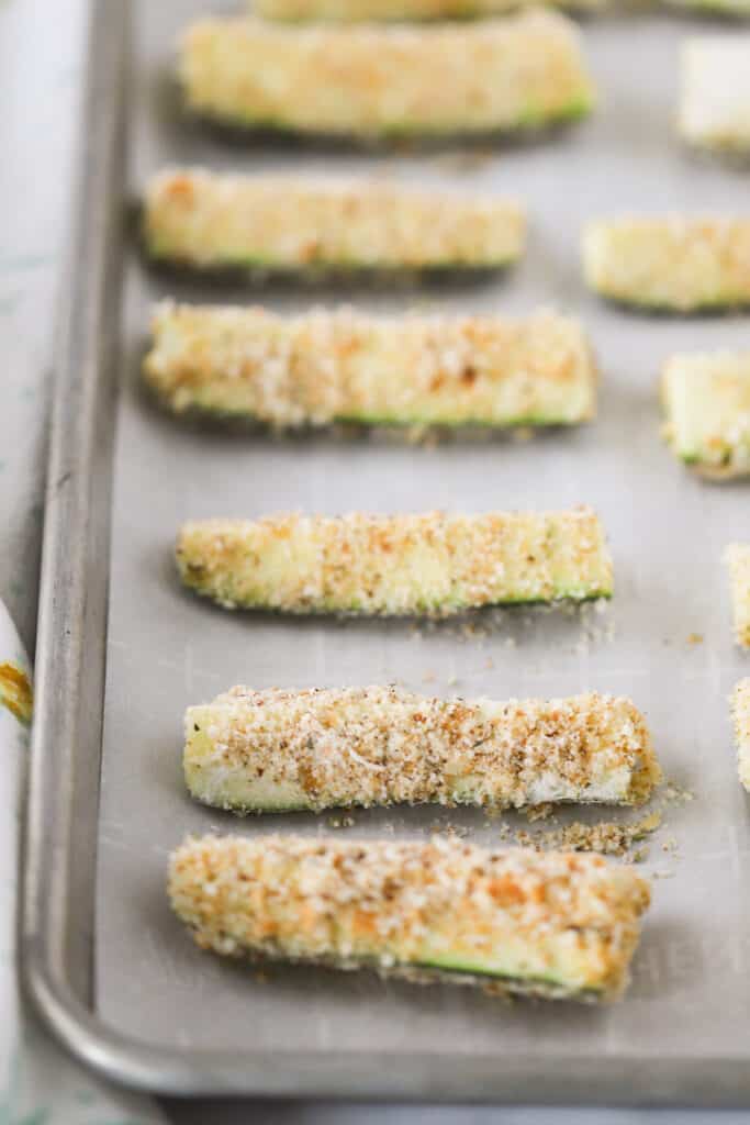 A sheet tray lined with parchment paper that has zucchini sticks lined up.  They are covered in a bread crumb mixture and ready to bake in the oven. Best zucchini fries recipe.