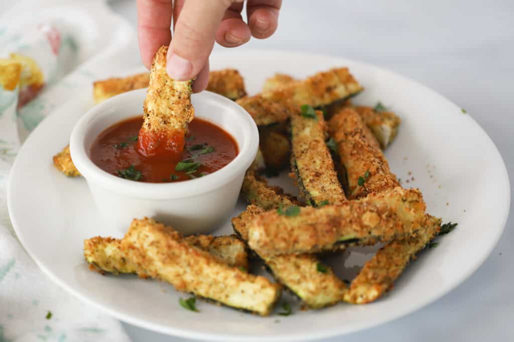 A serving plate full of baked Zucchini Fries with a side ramekin of marinara sauce.  A hand is holding a fry and dipping it into the sauce.