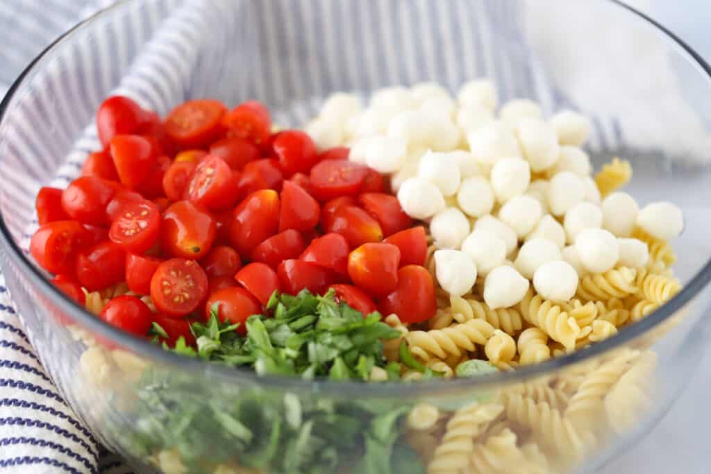 A glass mixing bowl filled with ingredients to make this Caprese Pasta Salad recipe, including cooked pasta, mozzarella balls, cherry tomatoes and fresh basil.