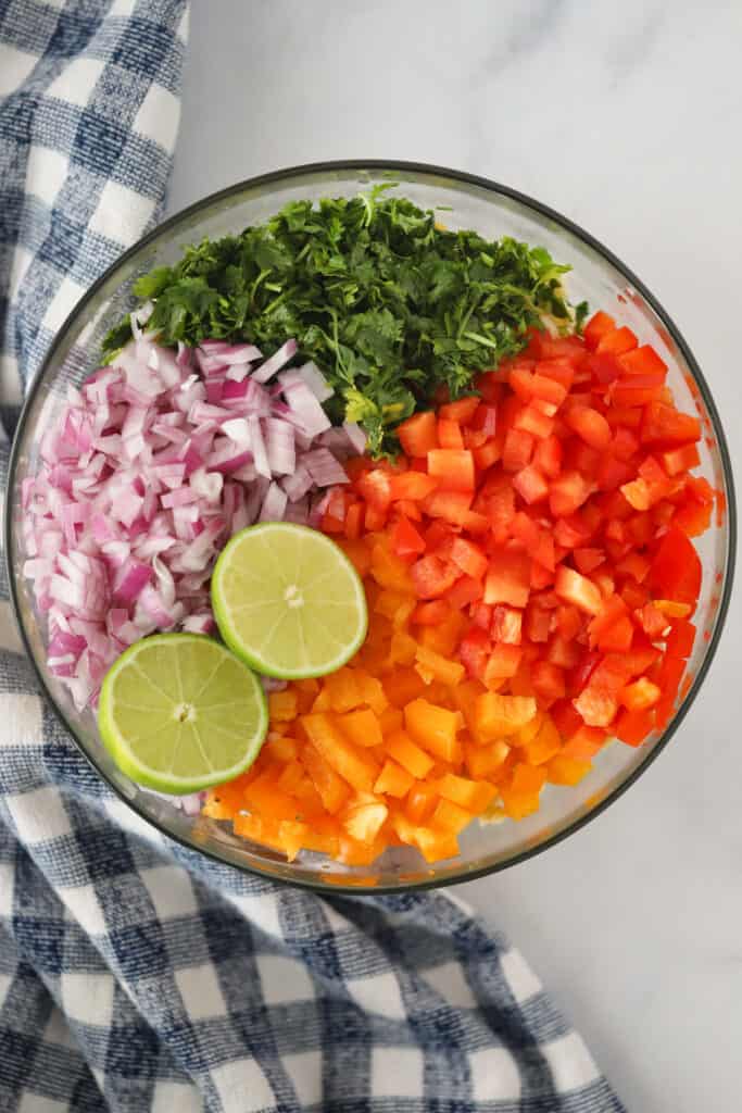 A mixing bowl with chopped red onions, cilantro, bell peppers and a sliced lime ready to be mixed.
