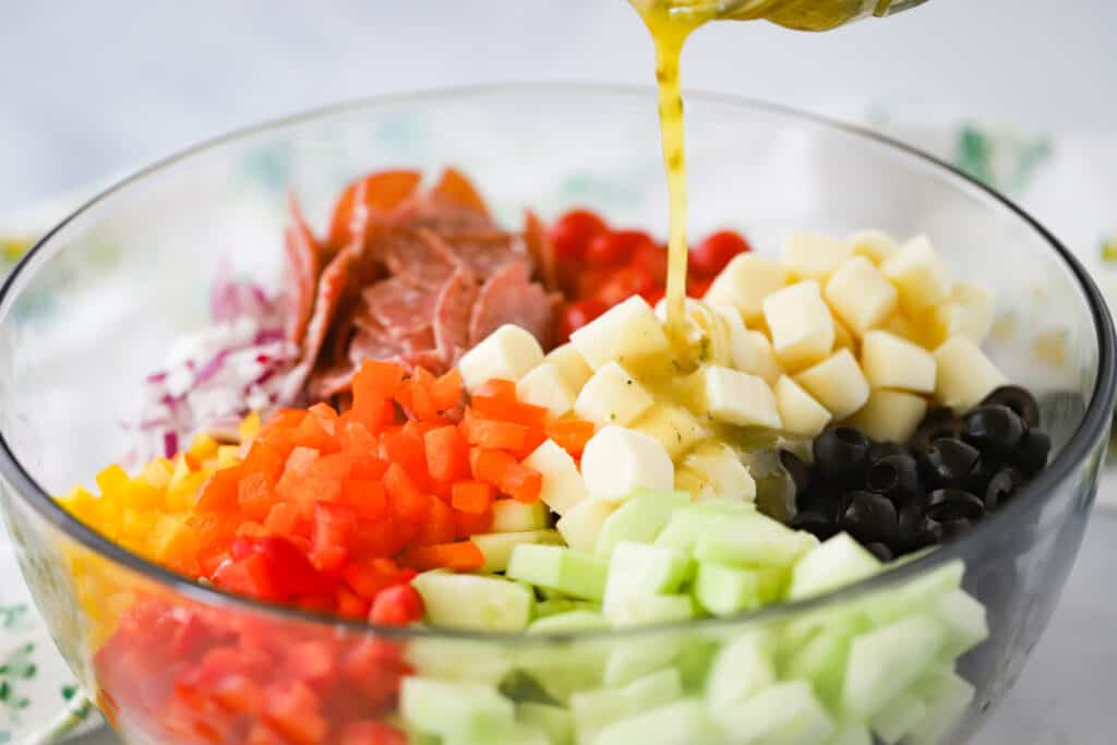 A bowl full of Italian pasta salad ingredients with salad dressing being poured in.