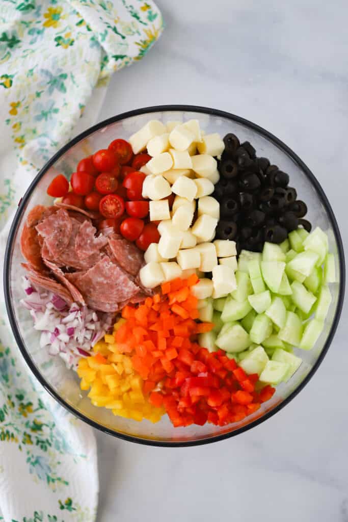 A glass mixing bowl filled with ingredients for making spiral pasta salad, including tomatoes, salami, onions, peppers, cucumbers, olives and cheese. 