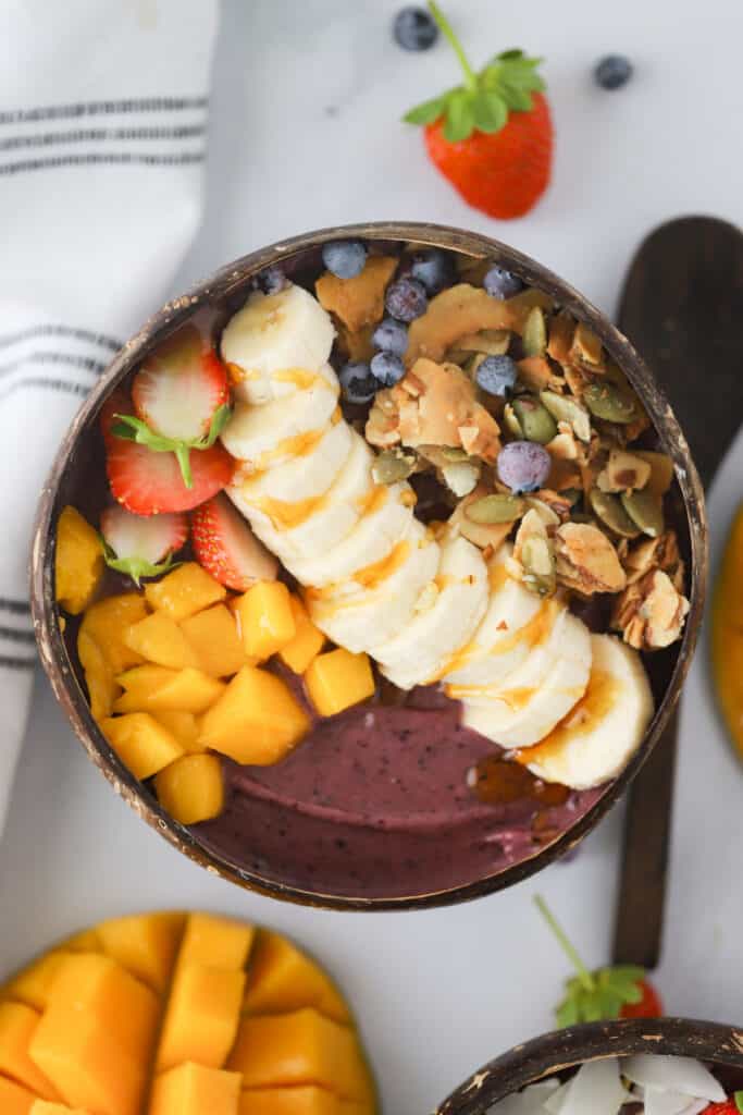 A homemade Acai Bowl on a tabletop, topped with mangos, strawberries, strawberries, granola, blueberries and honey.