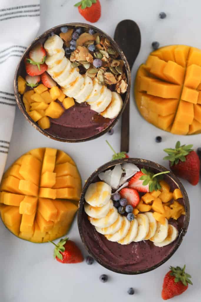 A tabletop with two Acai Bowls recipe topped with sliced bananas, mangoes, strawberries and granola.  Fresh mangos, blueberries and strawberries are also on the table. homemade acai bowl. 